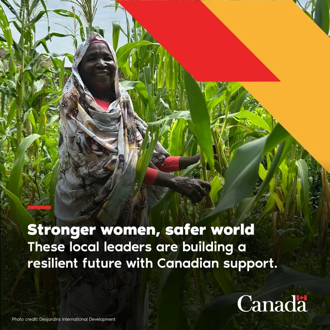 A woman stands in a field of tall green corn plants under a cloudy sky. She is holding one of the stalks and smiling.