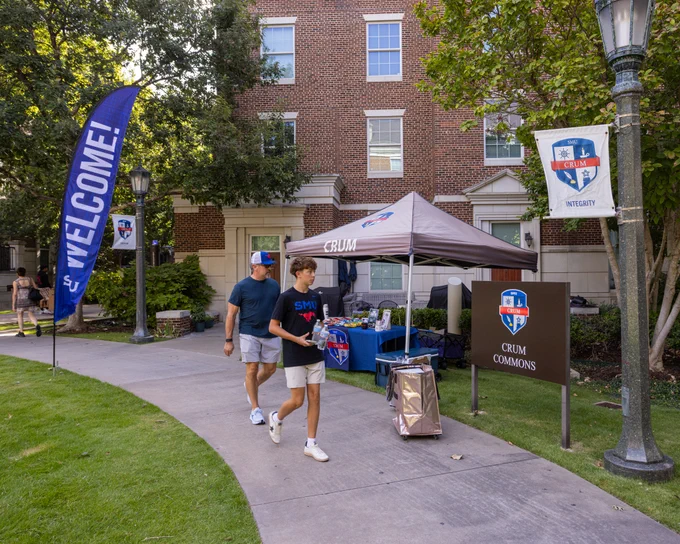 A person walks with another by a welcome booth outside a brick building labeled "Crum Commons." The booth has a tent with "Crum" on it, surrounded by banners and a sign with a crest and the word "Integrity." Trees and grass line the sidewalk.