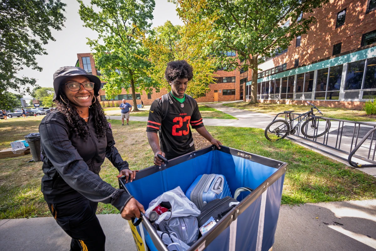Two people are pushing a large blue cart filled with personal items outdoors heading into Allyn hall at Kent State. 