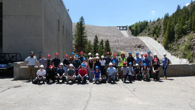A group of approximately 40 people in hard hats, mostly college students, poses in front of a power plant and spillway.