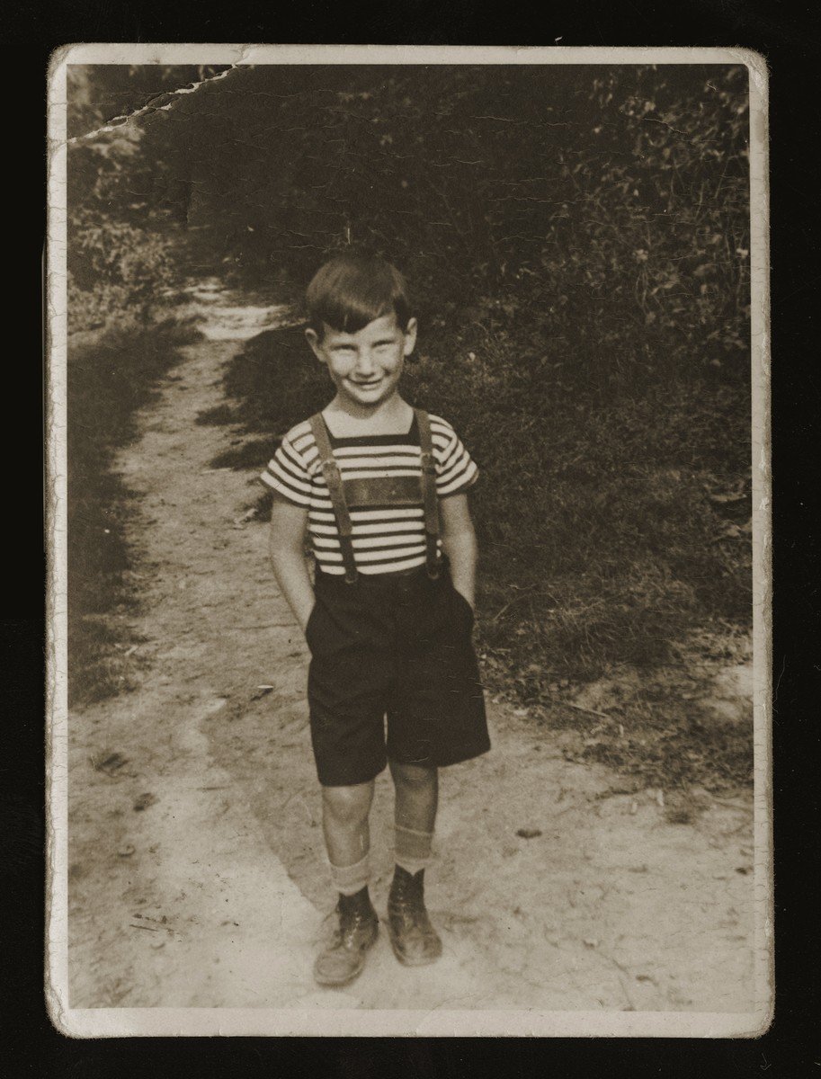 Black and white photo of a smiling child wearing a striped shirt and suspenders, standing on a dirt path.