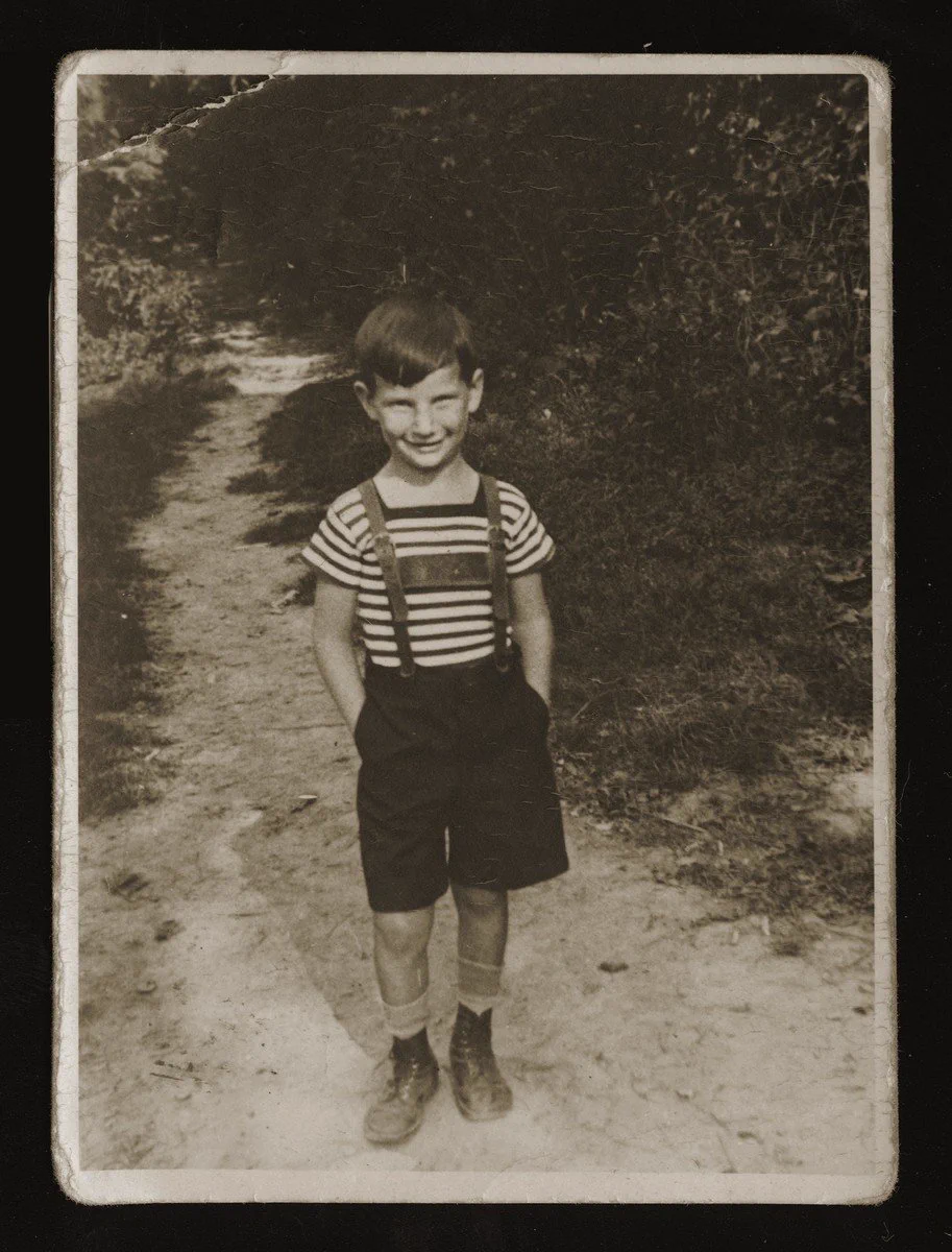 Black and white photo of a smiling child wearing a striped shirt and suspenders, standing on a dirt path.