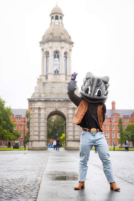 SuperFrog poses energetically in front of the Campanile at Trinity College Dublin. The background features an overcast sky and historic buildings.