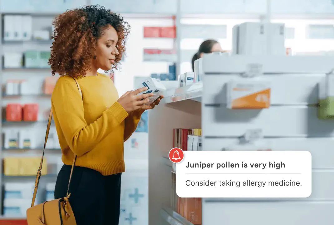A person with curly brown hair and a yellow sweater stands in a pharmacy aisle, carefully reading the label of a small white box of medication. In the foreground, a digital notification overlay features a red bell icon and reads, "Juniper pollen is very high. Consider taking allergy medicine."