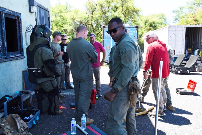 A group of people in tactical gear stand and converse outside near a building. Equipment and various tools are scattered on the ground. There are trees and storage containers in the background. Water bottles and a red toolbox are visible.