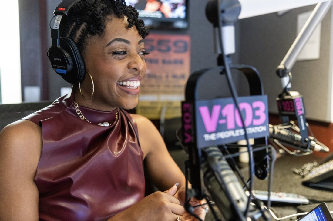 A person is sitting in a radio studio wearing headphones, smiling while speaking into a microphone. A sign for V-103, a radio station, is visible in the foreground.