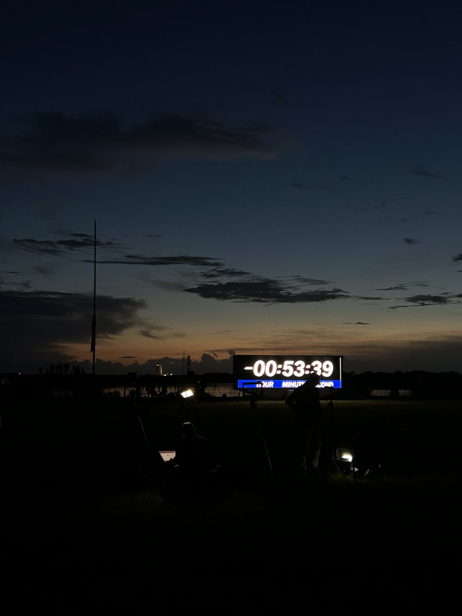 A massive rectangular screen lit up with a countdown clock that reads “-00:53:39” against a very early sunrise. The foreground is dark.