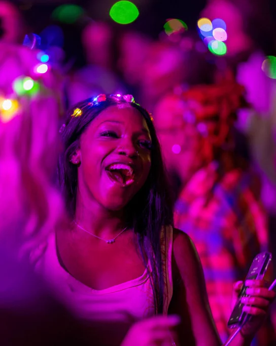 An individual is joyfully singing at a lively party, surrounded by colorful lights and blurred people in the background. They hold a phone in one hand and wear a light-up accessory on their head.