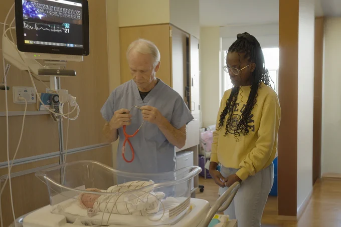 A healthcare professional in scrubs holds a stethoscope near a hospital bassinet containing a newborn. Another person stands nearby, observing. A medical monitor is visible, displaying vital signs. The setting is a hospital room.