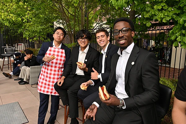 Four individuals in formal attire are seated outdoors, enjoying hot dogs. One person wears a red and white checkered apron, suggesting involvement in serving or cooking food. Lush green trees and a wrought iron fence provide a relaxed backdrop.