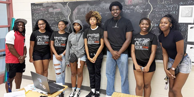 Group of eight diverse young adults standing in front of a classroom chalkboard, participating in the STAND summer program at the Homewood Center. 