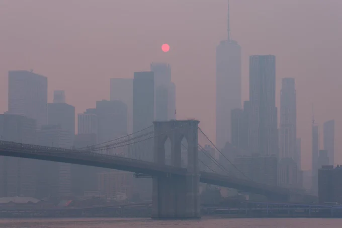 A hazy cityscape of New York City with the Brooklyn Bridge in the foreground. The skyline includes the One World Trade Center, and a reddish sun is visible through the smog.