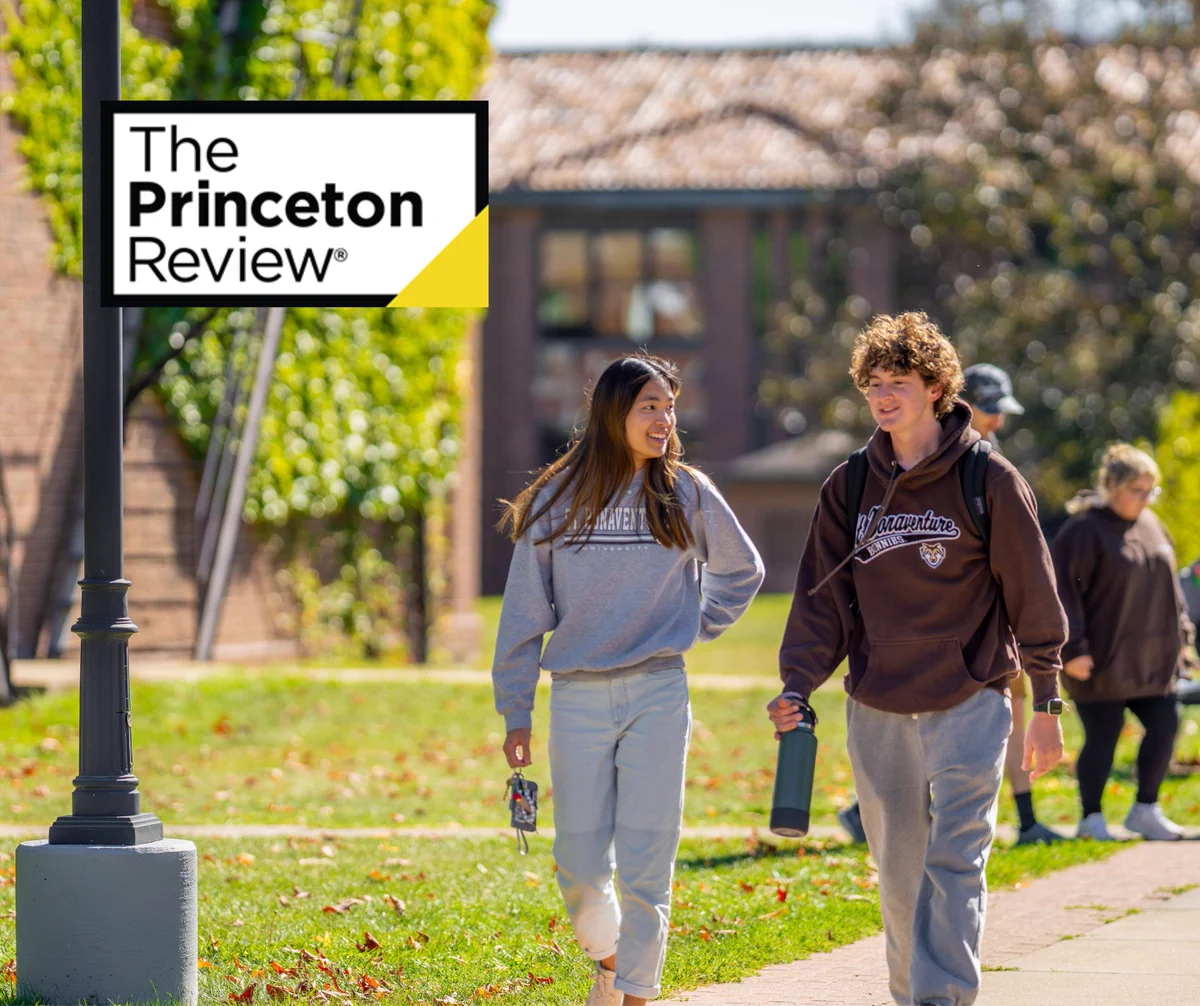 Two college students walk and chat on a sunny campus path with green grass and trees around them.