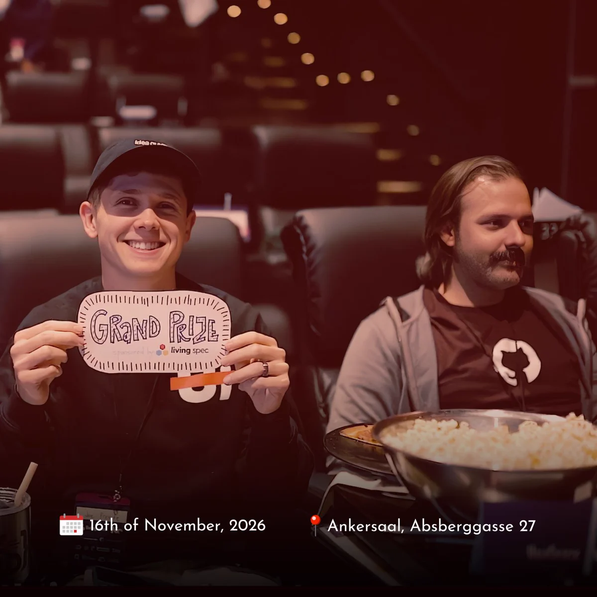 Two HalfStack attendees sit in an auditorium, with one smiling and holding a handmade “Grand Prize” sign. A large bowl of popcorn sits in the foreground. The graphic includes the HalfStack Vienna 2026 event details: 16 November 2026 at Ankersaal, Absberggasse 27.