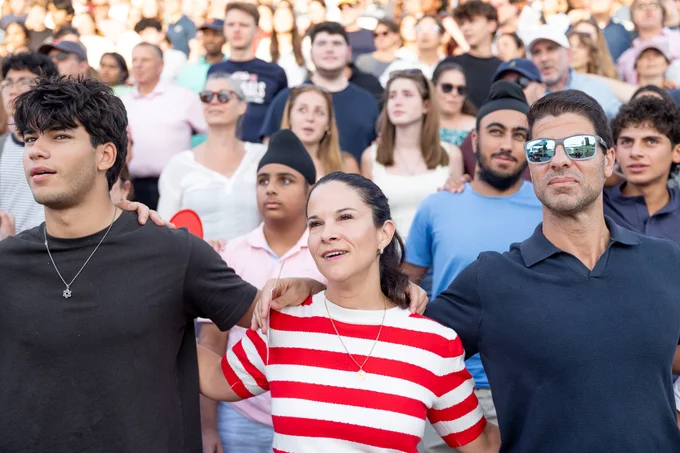 A group of people standing closely together in a stadium-like setting, appearing engaged with the event they are watching. Among the crowd, several individuals have arms around each other's shoulders, creating a sense of camaraderie.