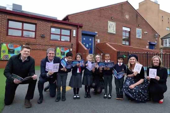 A group of adults and children kneel and stand in front of a school building. They are holding and displaying books, smiling towards the camera. The school has brick walls with colorful artwork on a nearby fence.