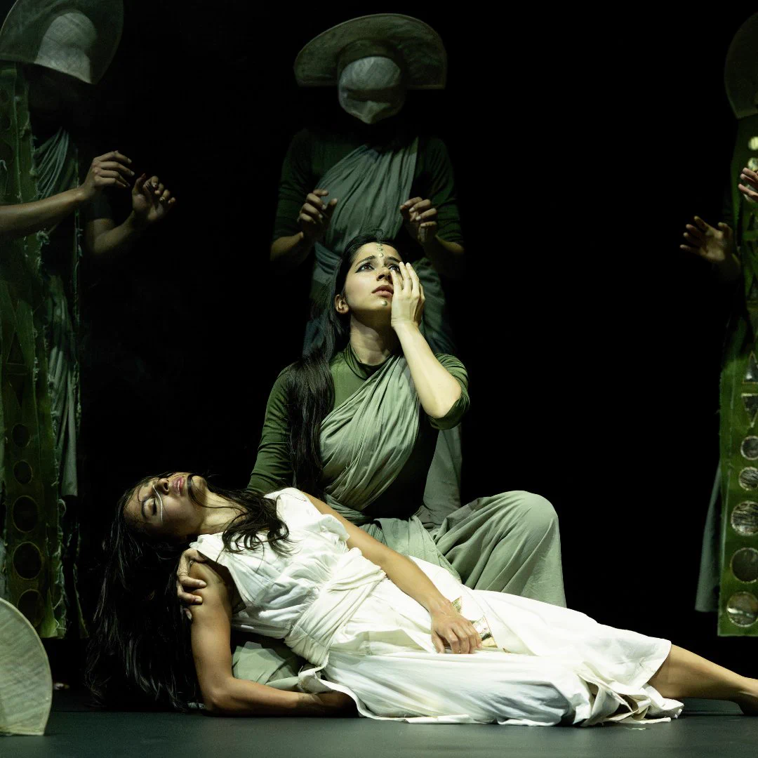 Image descriptions: 4 image carousel. The first are two dancers, one laying on the floor in the lap of a dancer who is seated and looking up towards the sky. Masked dancers stand behind them. The second is a dancer wearing red posing with their hands out in front of them. The third is a dancer striking a pose, holding up fabric behind their head. The fourth is a dancer wearing white, seated on the ground, with a large rock in front of them.