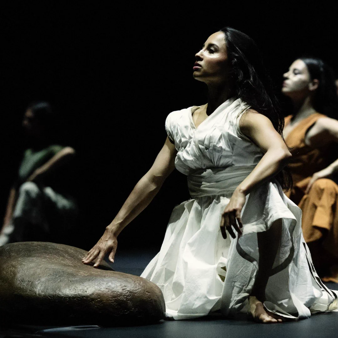 Image descriptions: 4 image carousel. The first are two dancers, one laying on the floor in the lap of a dancer who is seated and looking up towards the sky. Masked dancers stand behind them. The second is a dancer wearing red posing with their hands out in front of them. The third is a dancer striking a pose, holding up fabric behind their head. The fourth is a dancer wearing white, seated on the ground, with a large rock in front of them.