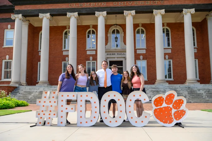 President Ayers posing for a photo with a group of students with the FDOC letters in front of Sikes Hall.