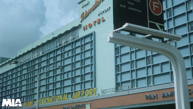 Low-angle shot of the retro Miami International Airport Hotel exterior featuring blue grid paneling, vintage orange signage, and street light posts under a cloudy sky. The MIA logo is in the bottom-left corner.