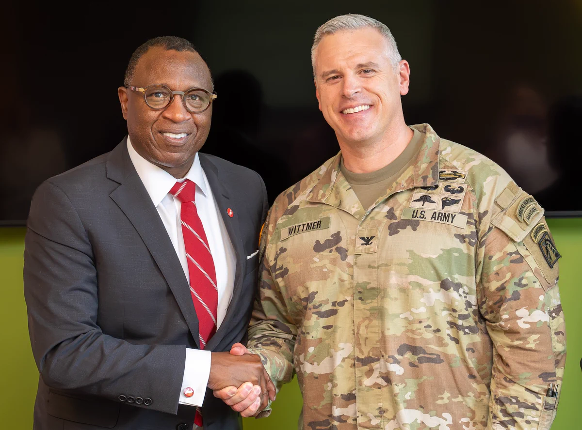 Chancellor Howell in a dark suit with a red tie and glasses shaking hands with a man in a U.S. Army uniform smiling warmly.