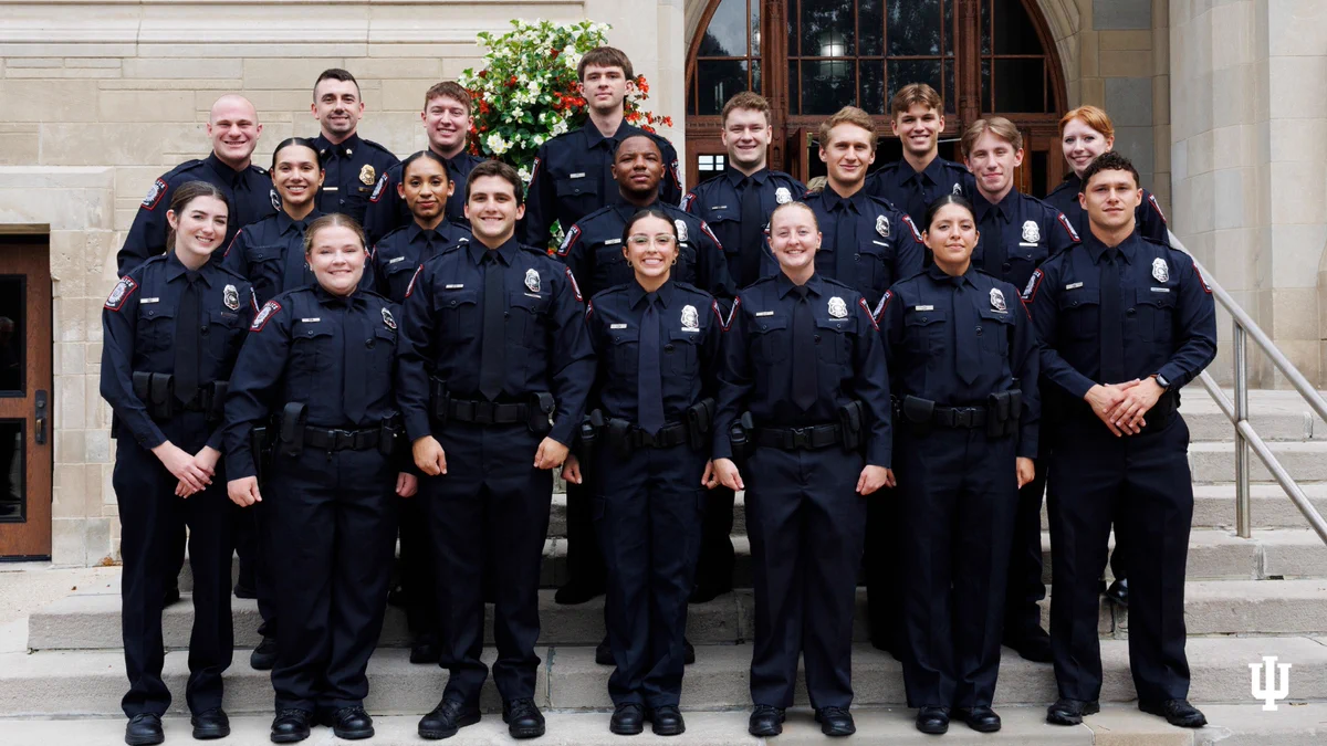 A group of uniformed individuals stands on the steps of a building with stone walls. They are wearing dark uniforms with badges. The group appears to be neatly arranged in rows, and they are standing in front of a doorway. The building seems institutional, and the Indiana University (IU) logo is visible in the corner.