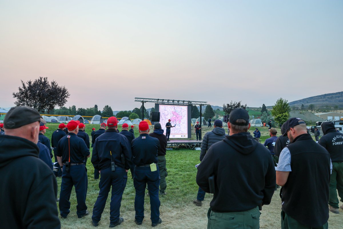 Firefighters stand in an open field listening to a person delivering a briefing on a stage with a map behind them. Tents are in the background.