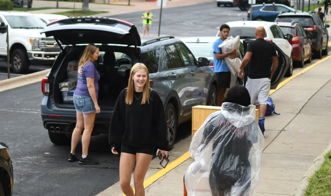 A group of people is engaging in moving activities on a sidewalk adjacent to parked cars. Some are carrying boxes and bags. A woman in a purple shirt stands near an open vehicle trunk filled with belongings. A person in a rain poncho walks away from the camera. The atmosphere suggests an organized event, possibly a move-in day.
