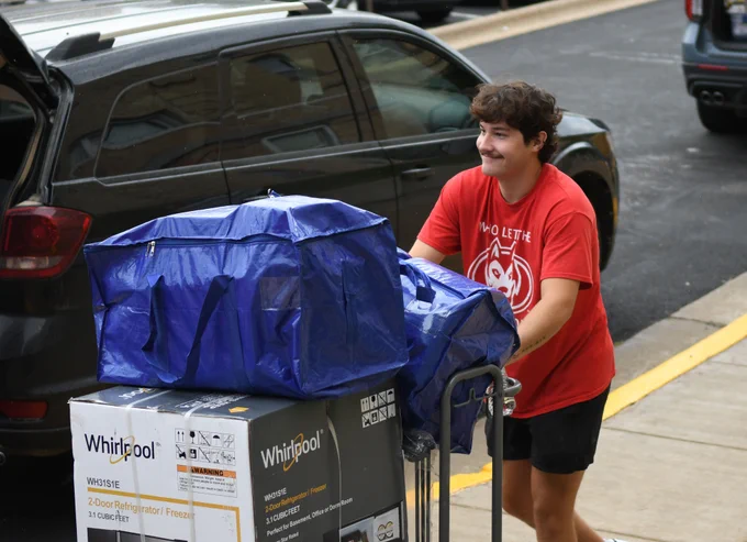 A person wearing a red shirt with a logo is pushing a cart loaded with large boxes. The boxes display the Whirlpool brand. They appear to be unloading from a dark-colored vehicle parked on the street.