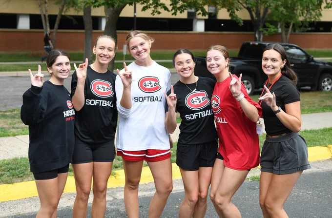 A group of six people is standing outdoors, smiling and gesturing with their hands. They are wearing casual athletic clothing with "Huskies Softball" logos. Trees and parked cars are visible in the background.
