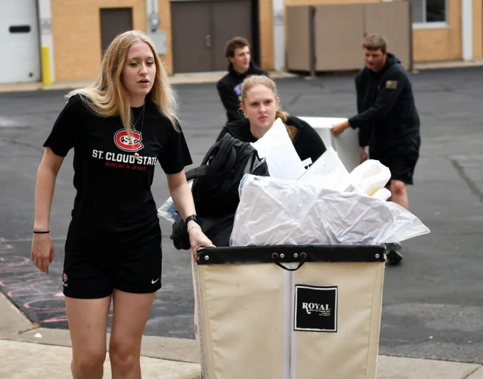 A group of people, some wearing St. Cloud State University apparel, are pushing and pulling carts filled with personal belongings outdoors, possibly moving items into or out of a building. The background shows an urban environment with brick walls and doors.