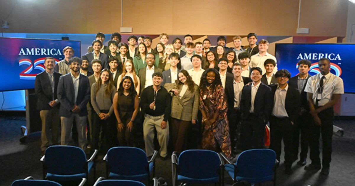 A large group of people standing and posing in a studio setting with two screens displaying "America 250" in the background. The group is composed of individuals with a variety of appearances, dressed in formal and semi‑formal attire. The setting suggests an event or gathering, with chairs visible in the foreground.