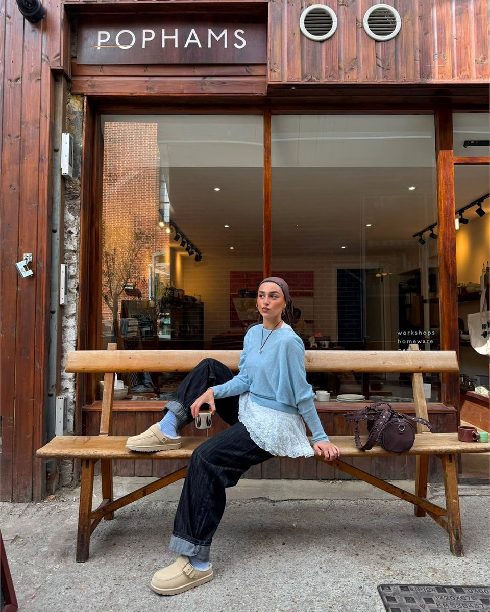An image of a person wearing a pair of sand toned UGG Goldenstar Evelina Clogs with denim jeans, blue top and brown headscarf whilst sitting on a wooden bench outside a cafe.