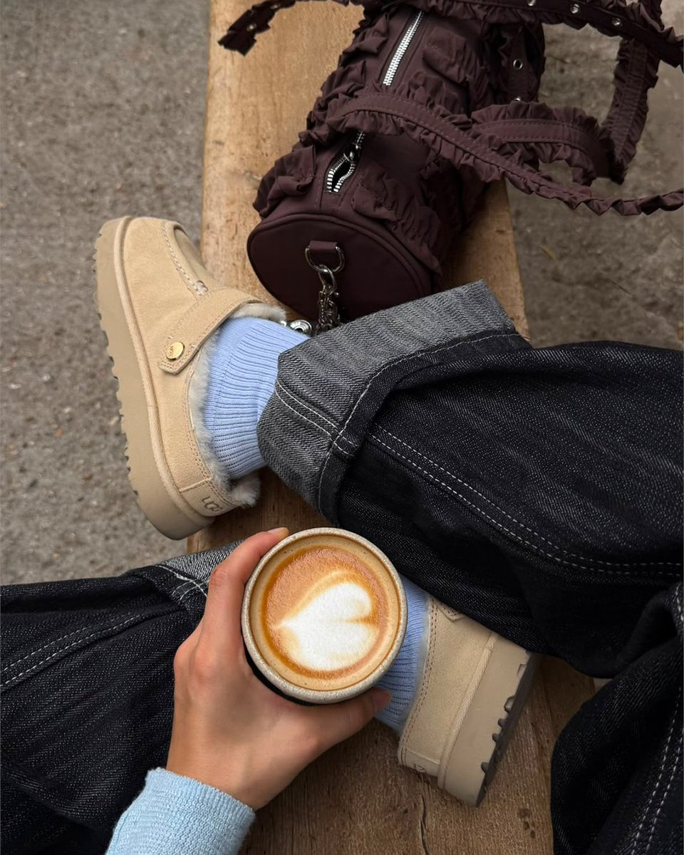 An image of a person's lower legs wearing a pair of sand toned UGG Goldenstar Evelina Clogs with denim jeans whilst sitting on a wooden bench.