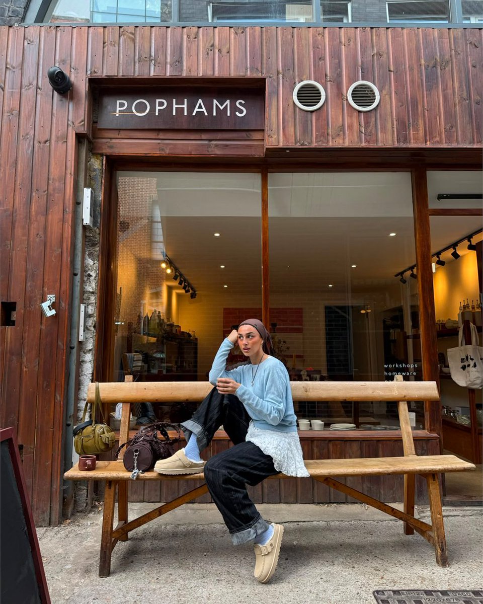 An image of a person wearing a pair of sand toned UGG Goldenstar Evelina Clogs with denim jeans, blue top and brown headscarf whilst sitting on a wooden bench outside a cafe.