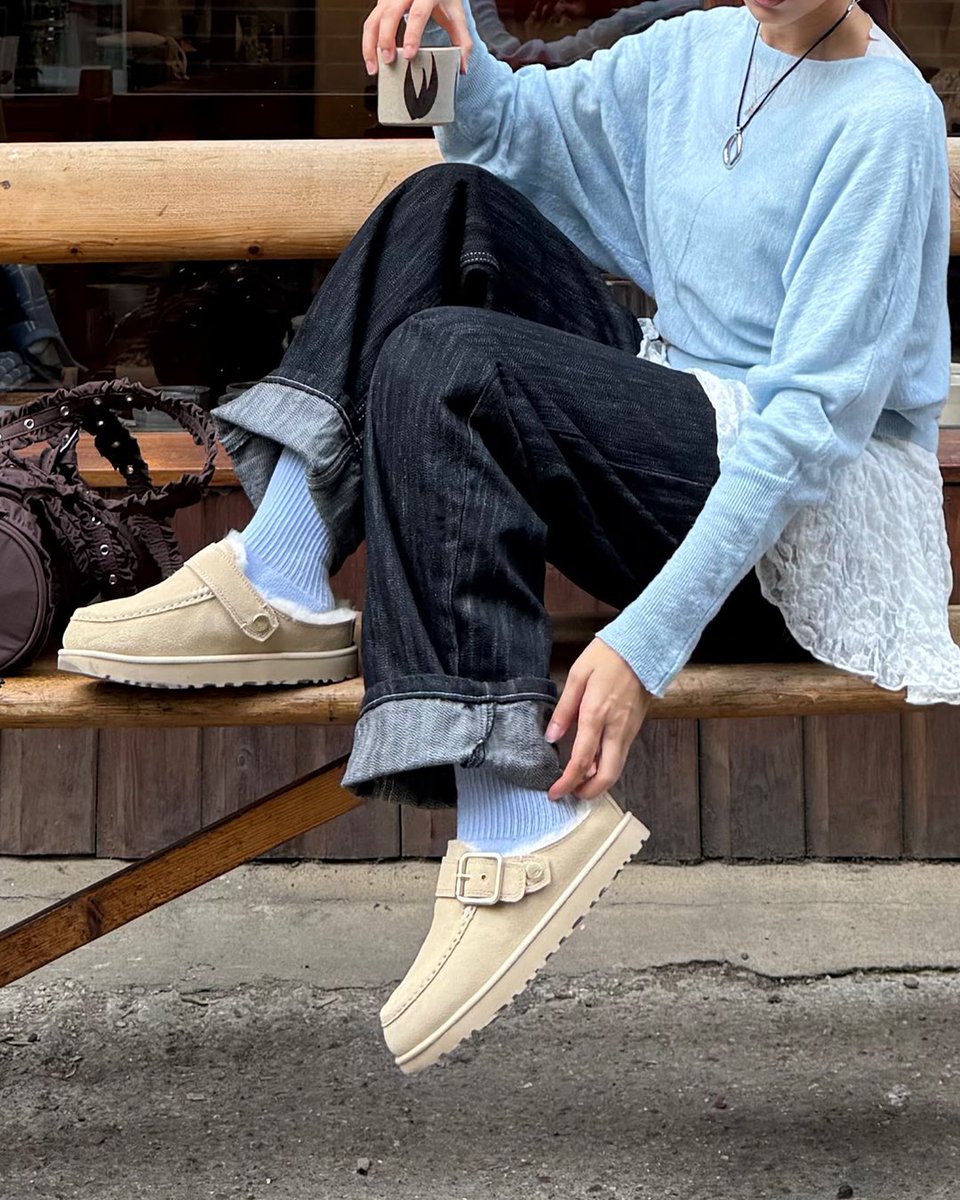 An image of a person wearing a pair of sand toned UGG Goldenstar Evelina Clogs with denim jeans, blue top whilst sitting on a wooden bench outside a cafe.