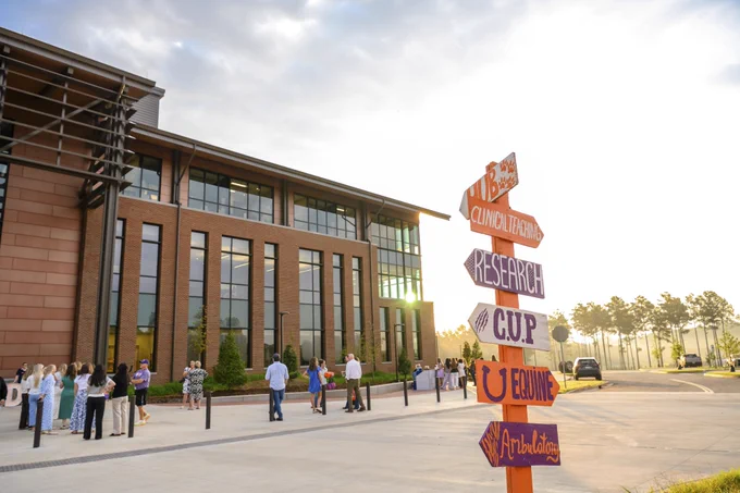 An outdoor scene featuring a modern building with large glass windows. A colorful signpost in the foreground points to various locations, including "Clinical Trials," "Research," and "Equine." Several people are standing nearby on a sunny day.