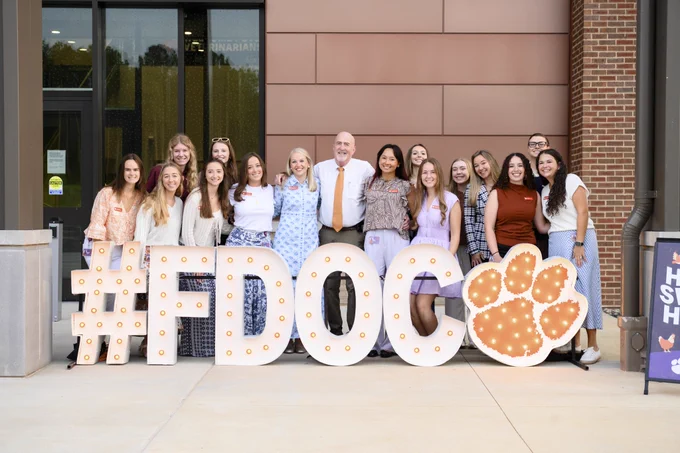 A group of people is standing in front of a large illuminated "#FDOC" sign with the Clemson Tiger Paw print design. They are smiling and gathered closely together outside a college building's entrance.