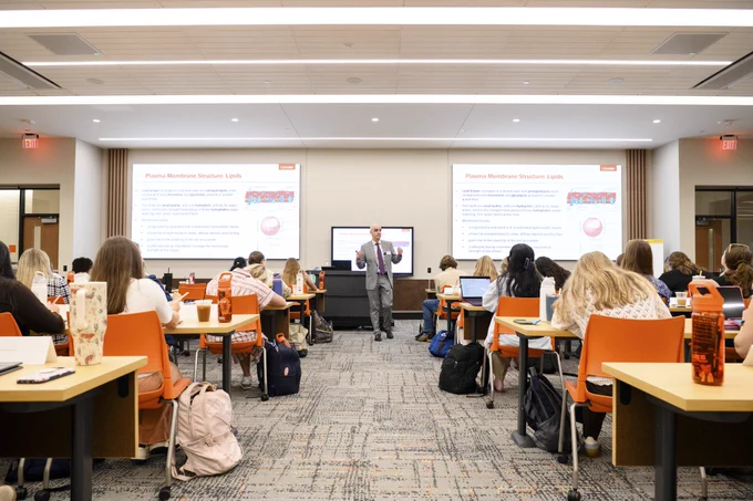 A classroom filled with students sitting at desks facing a presentation screen. An instructor is standing at the front, giving a lecture. The presentation on screen displays the topic "Plasma Membrane Structure: Lipids," with text and diagrams.