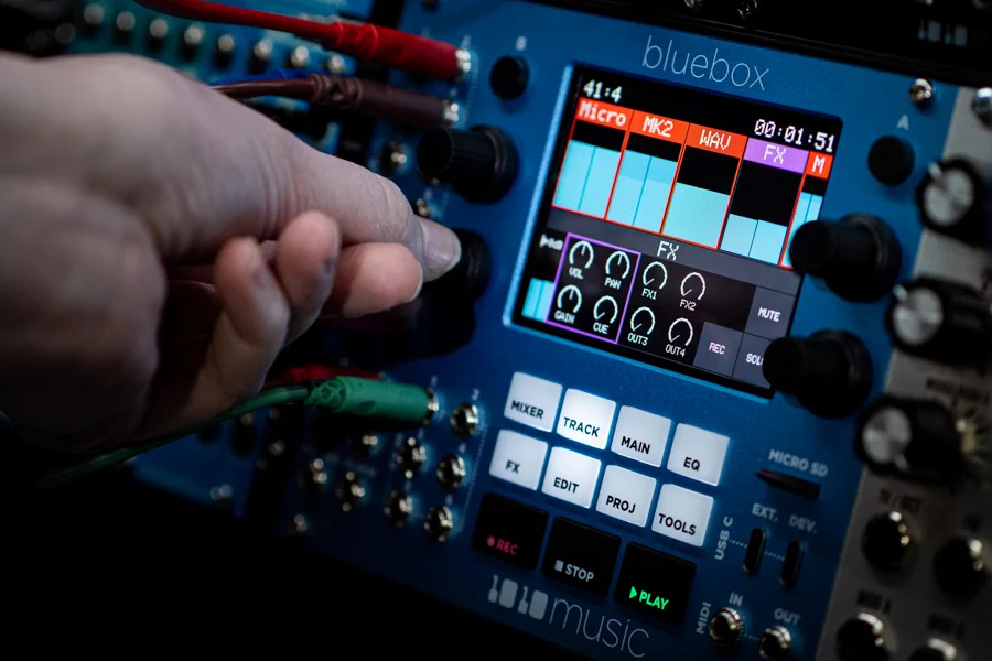 A hand reaches out and touches a Bluebox Eurorack module, color touchscreen showing channels and FX bus, FX parameters, button shortcuts, transport, and a couple of cables connected to I/O minijack, microSD slot, etc.
