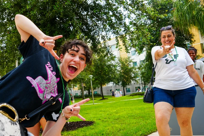 Two people pose outdoors with lush greenery and trees around them. One person is wearing a black shirt with a vibrant graphic and is giving the "horns up" hand sign. The other in a white shirt has a USF logo and is gesturing cheerfully.