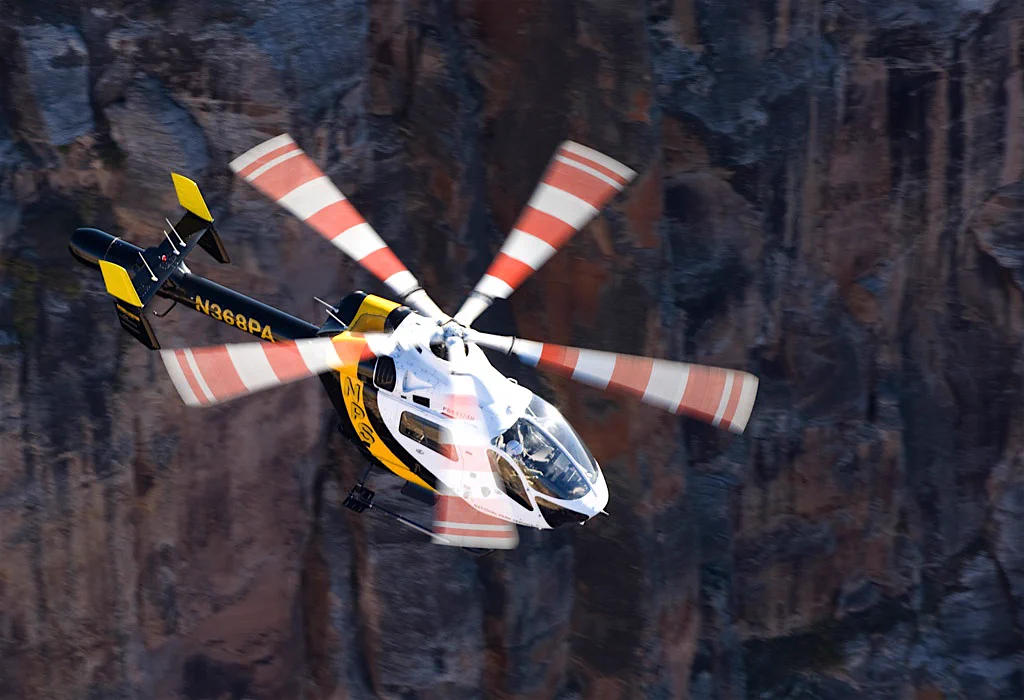 White and yellow helicopter with spinning red-striped rotor blades flying near a rugged cliff face.