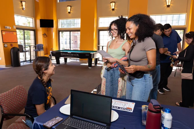 A person is seated at a table with a laptop and information sheets, speaking with two others holding flyers. Behind them, there is a Brunswick pool table. The setting is at one of UTEP's residence halls in an indoor space with large windows and several people engaged in conversation.