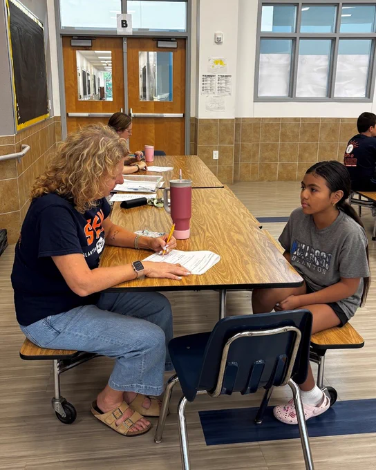 Two people, a woman and a girl, sitting at a table in what appears to be a school or classroom setting. The woman has curly hair and is wearing a navy shirt, while the girl has short dark hair and is wearing a gray shirt.