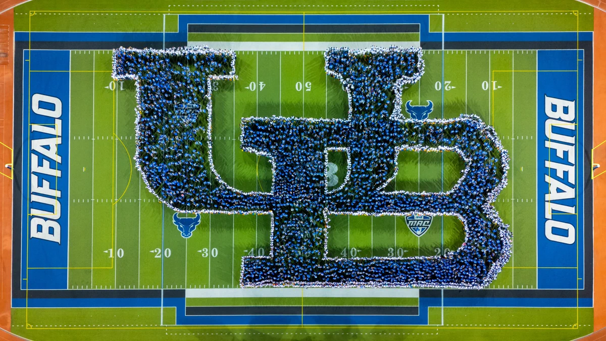 Aerial view of hundreds of people in blue and white shirts forming large 'UB' letters on a green football field. \"BUFFALO\" is written in white on blue end zones, with blue bull and '1946 MAC' logos also visible on the turf.
