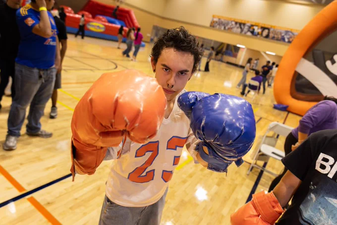 A person wearing oversized orange and blue boxing gloves is playfully posing at an indoor event. The setting includes inflatable attractions like a slide and various attendees in the background. The overall atmosphere is lively and energetic.
