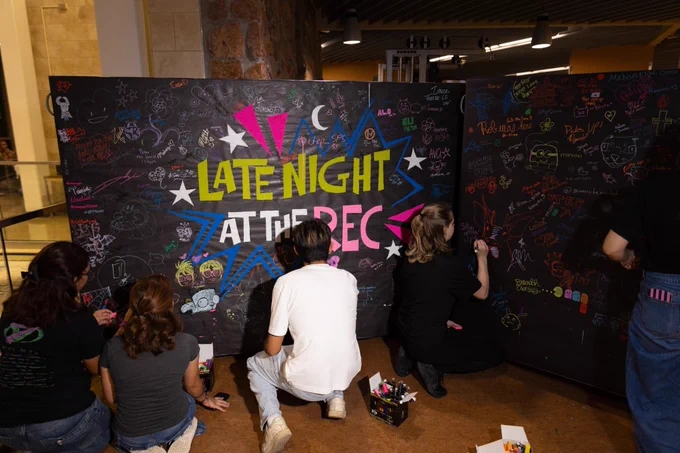 People are writing on a large blackboard with colorful chalk at an event titled "Late Night at the Rec." The board is filled with various drawings, messages, and decorations. Several people are crouched down, adding their contributions to the artwork.
