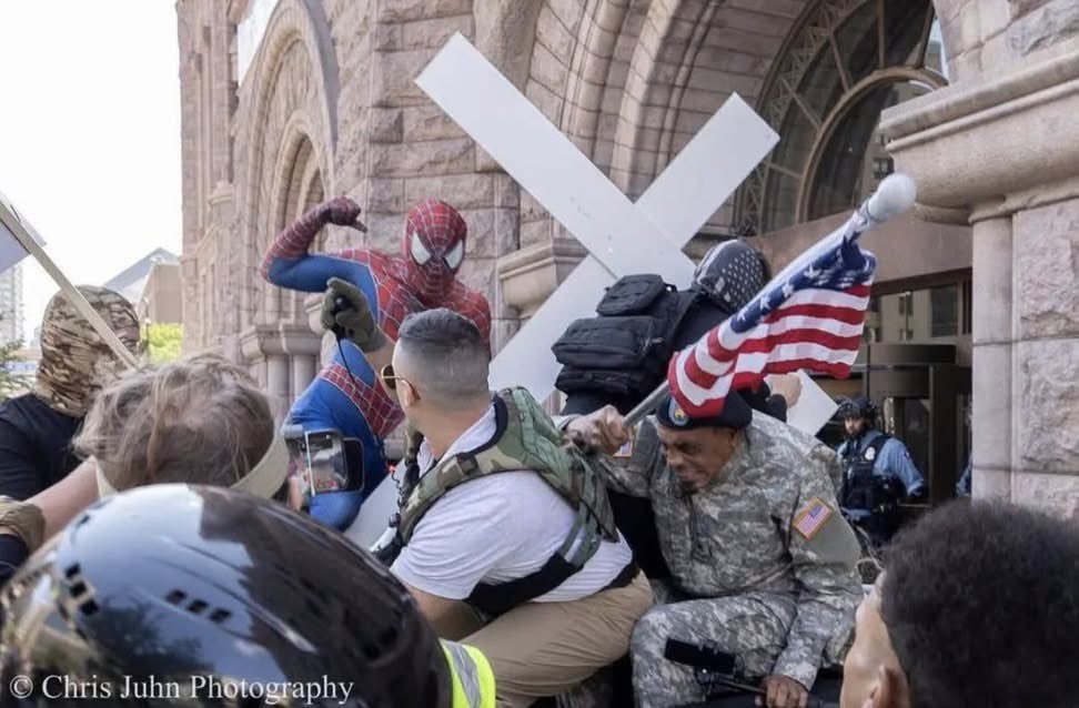 landpalestine's tweet photo. Peak 2026. The phone, the cross, the flag, the soldier, the cop. The American melee. The absurdity of Spidey in the midst of it. This photo is art https://t.co/NpVLXDqMJa