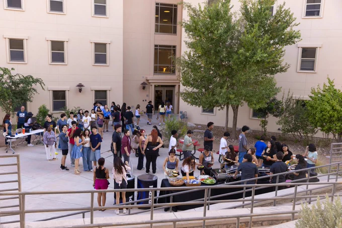 This image shows a social gathering in a courtyard  on the UTEP campus surrounded by a beige building with multiple windows. A diverse group of people mingles, some lining up at tables on the right where food is being served. Trees and greenery add a touch of nature to the concrete space.