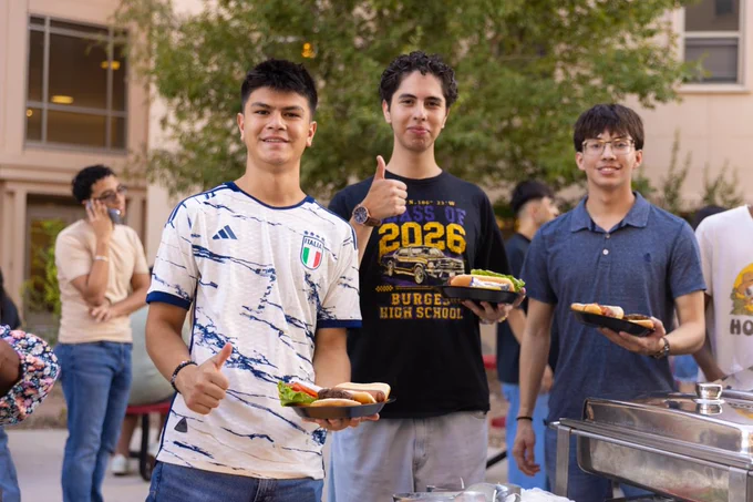 Three people are outdoors at a casual gathering, holding plates with burgers. One is wearing an Italy soccer jersey, another has a "Class of 2026" shirt from Burges High School, and the third is in a graphic t‑shirt. They are smiling, with some greenery and a building in the background.

